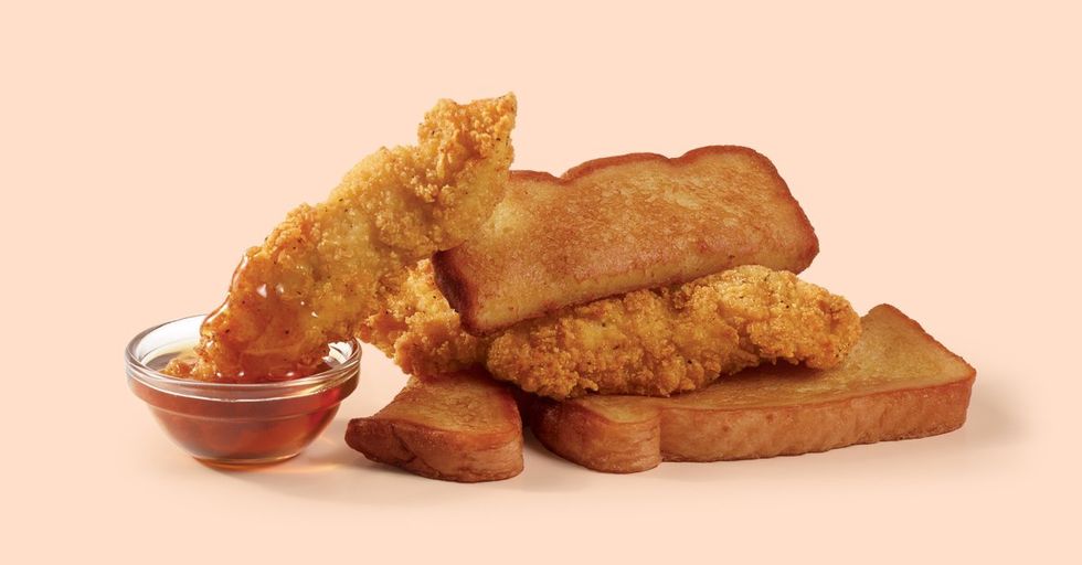 Three chicken strips and French toast sticks lay in a pile with one chicken strip angled to dip into a small glass bowl of syrup.