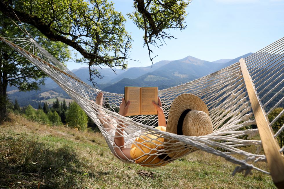 A person wearing a straw hat reads a book while laying in a hammock surrounded by nature.