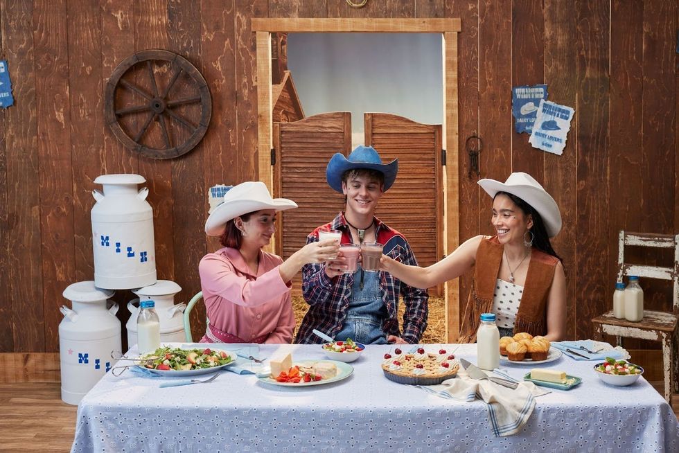 Three people cheers glasses of milk over a spread of delicious dairy foods in a barn.
