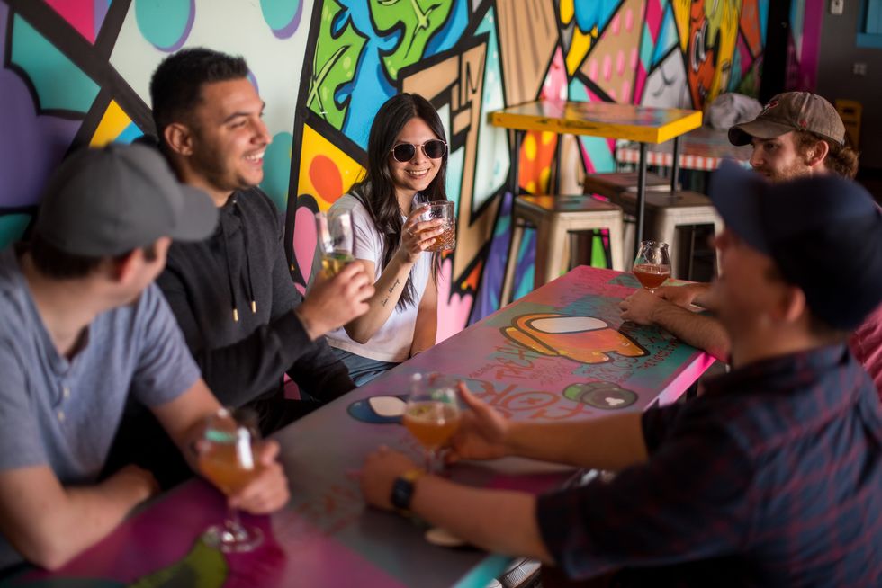 A group of friends sits around a colorful table inside a vibrant, mural-covered brewery, laughing and holding drinks while enjoying each other's company.