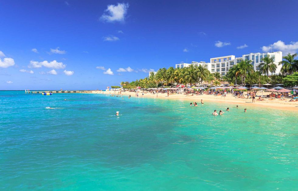 A beach with bright turqouise water, yellow sand, palm trees and a while hotel building.