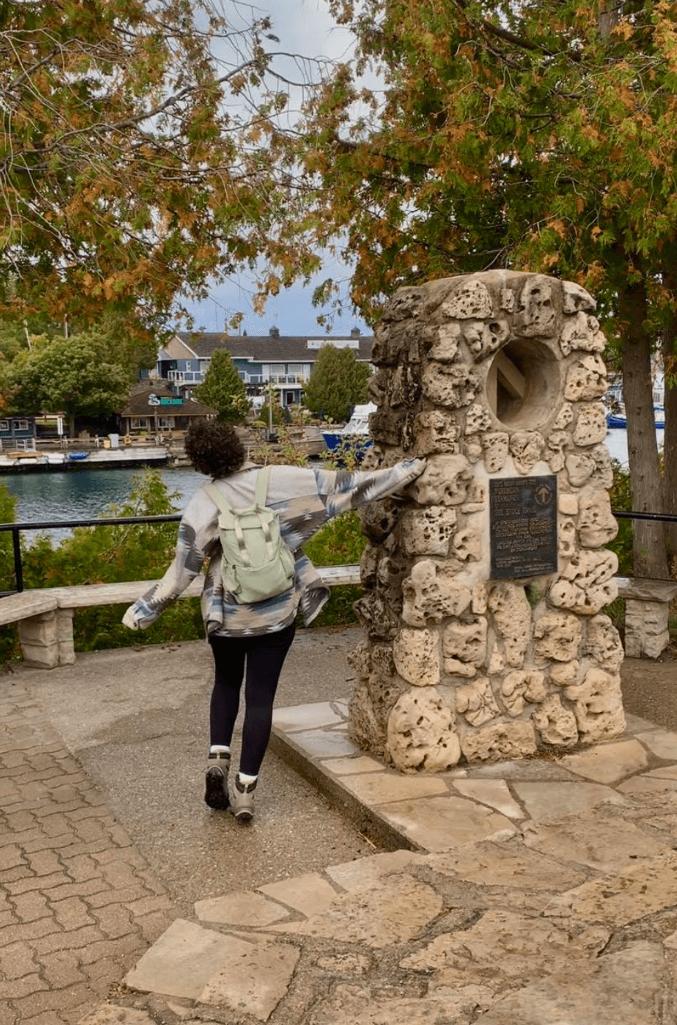 A person posing beside the stone Bruce Trail northern cairn overlooking Little Tub Harbour in Tobermory, Ontario, surrounded by autumn trees.